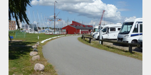 Zufahrt zu den Pltzen. Links im Bild ein kleiner Spielplatz, im roten Gebude sind Toiletten, Duschen, Hafenmeisterei sowie ein kleines Bcherregal untergebracht.